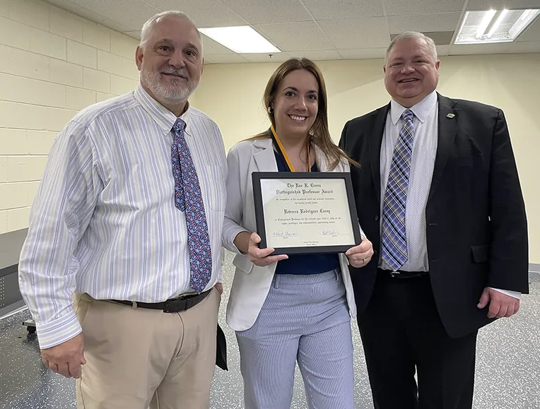 A woman holds a framed award certificate with two men in suitcoats on either side of her.