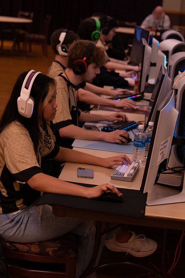 A woman and four men sit at tables in front of computer screens. They wear headphones and are concentrating on their electronic games.